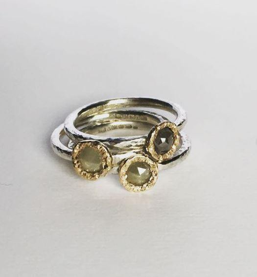 Three sterling silver stacking rings are stacked together. Each ring's band has a slightly hammered finish and is adorned with circular gems. The gems are set in golden bezels, featuring grey diamonds that appear to have a soft, translucent quality with a subtle sparkle. These exquisite pieces are known as the Aurous Ring Silver Grey Diamond by Abbie Dixon.