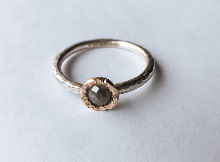 A close-up image of the Aurous Ring Silver Grey Diamond by Abbie Dixon, a delicate sterling silver stacking ring with a hammered finish on its band. It features a round, rough-cut grey diamond set in a slightly raised, textured bezel. The ring is placed against a plain, white background.