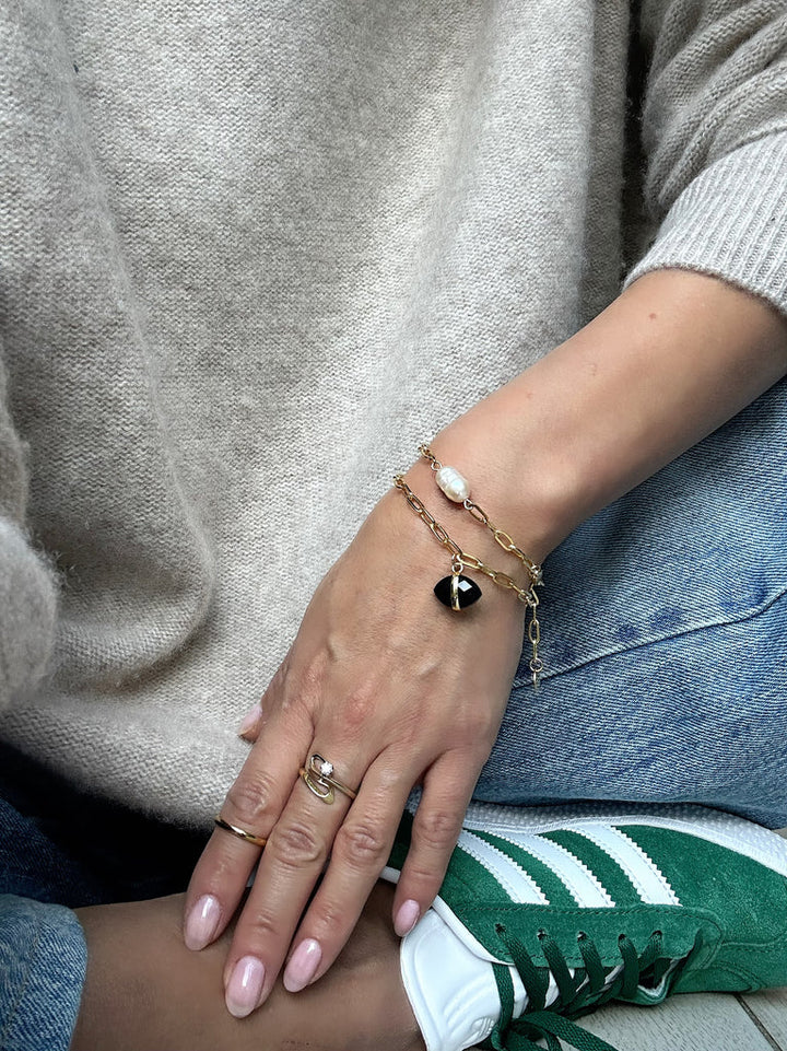 A person wearing a beige sweater and blue jeans with green and white sneakers is showing their left hand. They have a gold bracelet with a black charm and another bracelet with a white pearl on their wrist, complemented by the Ayin Bracelet in various gemstones from Aria V. Their nails are painted in a neutral shade.