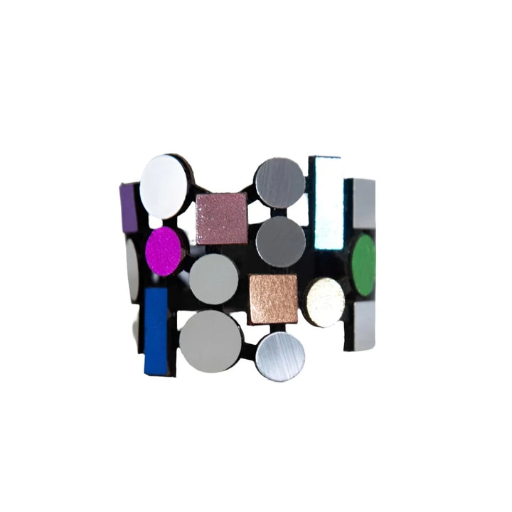 The Iskin Sisters' Bauhaus Bracelet, boasting a vibrant array of geometric shapes and finishes, lies elegantly on a stone-like textured surface. This striking piece features circles and rectangles in metallic, matte, and glossy hues including silver, purple, blue, green, black, and copper.