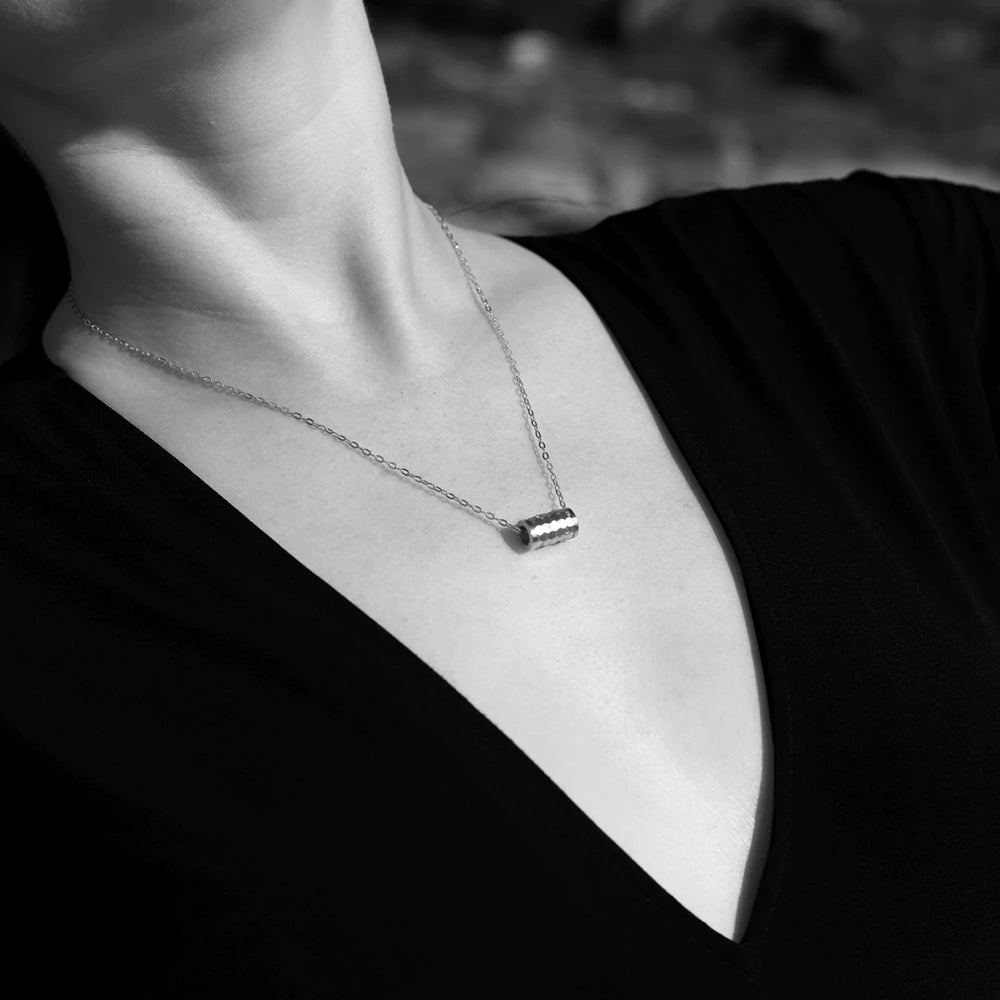 A person wearing a delicate Andrea Mears Jewellery Karma Wheel Necklace Silver featuring a small, cylindrical pendant. The person is dressed in a black, deep V-neck top. (Black and white image).