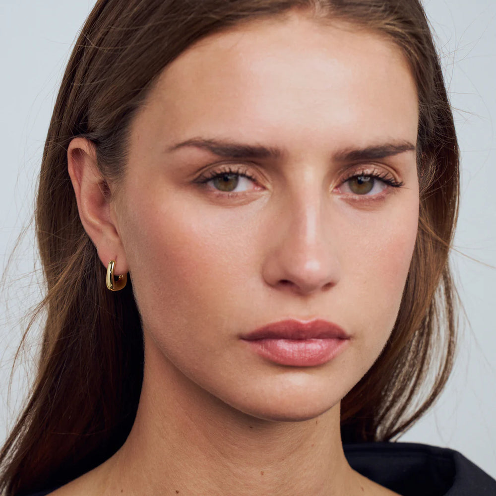A woman with light brown hair and minimal makeup models Sif Jacobs' Borsa Pianura Earrings—18k gold plated sterling silver—as she gazes neutrally at the camera. She wears a black top against a plain, light background.