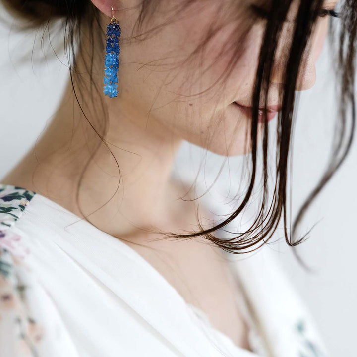 A woman with dark hair partially covering her face is shown in profile. She is wearing a white garment with delicate, floral patterns on the sleeves and a statement necklace from SAKURA Flower Jewelry. She has long, dangling Blue Sakura Earrings from Tochu-De and is looking down softly. The background is neutral and out of focus.