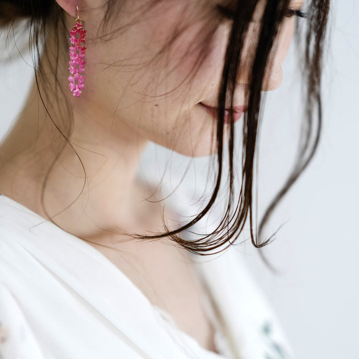 A close-up of a woman with dark hair wearing a white top. She is looking down, and her face is partially obscured by her hair. She wears a pair of Fuchsia Sakura Earrings from Tochu-De, which stand out against her light skin and white attire.