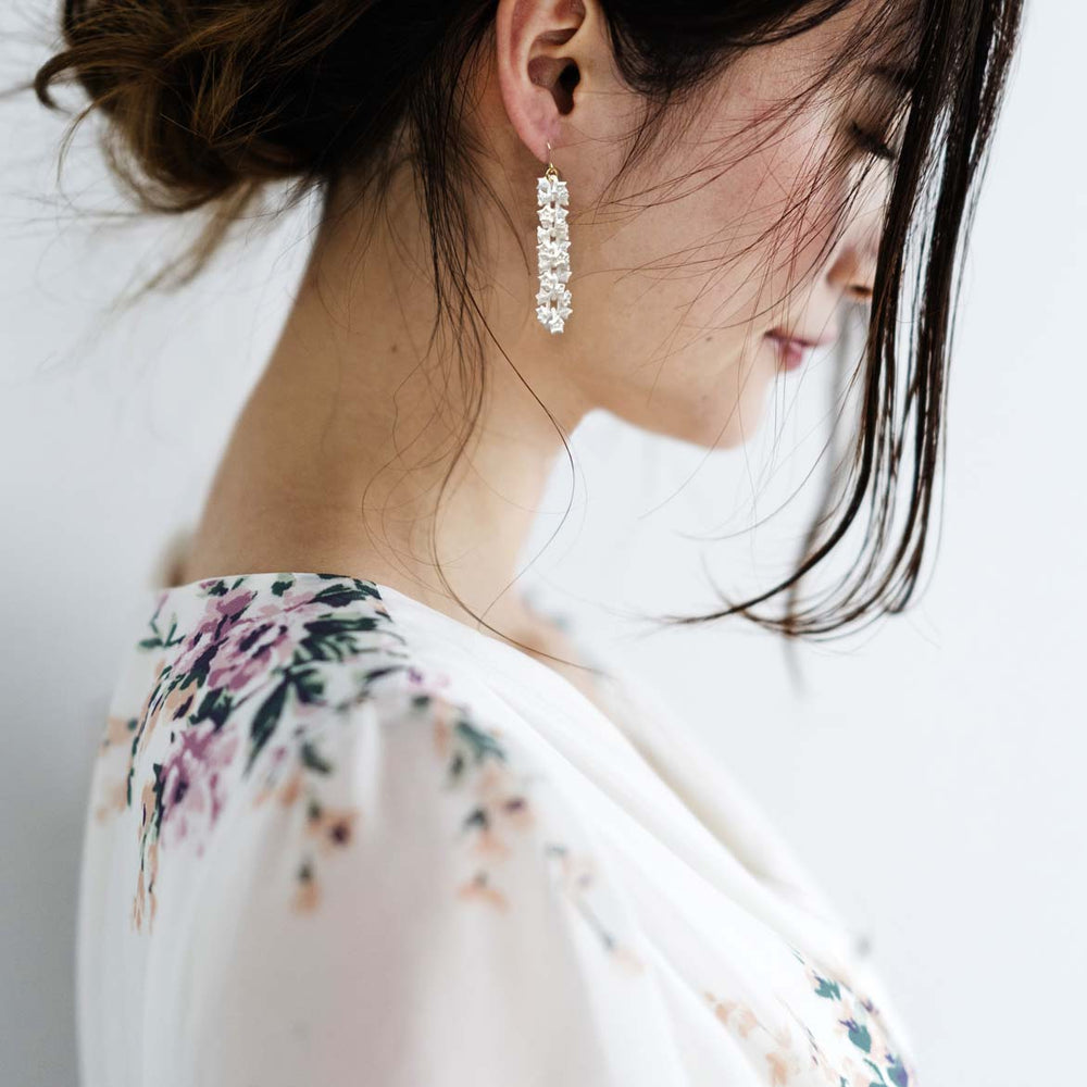 A woman with dark hair, tied back with a few loose strands framing her face, wears Tochu-De's long, dangling gold stainless steel White Sakura Earrings. She is dressed in a white floral robe or blouse and is looking downward, showing her profile against a plain light-colored background.