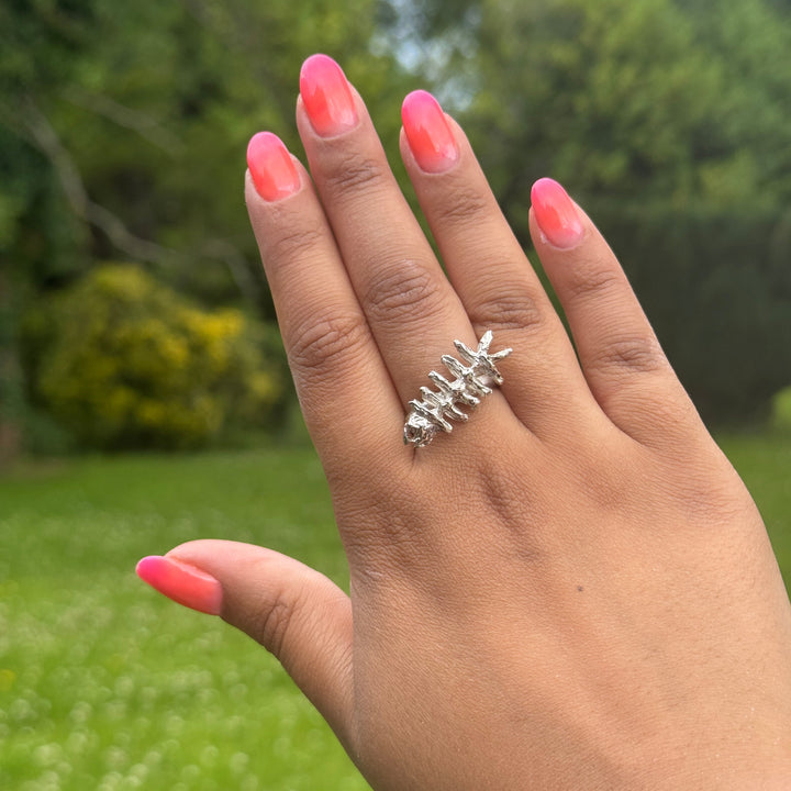A hand with pink ombré nails wears the Soufana Attia Baraka Ring, inspired by Arab symbols of protection and Baraka, shown outdoors against green grass with blurred trees and bushes in the background.