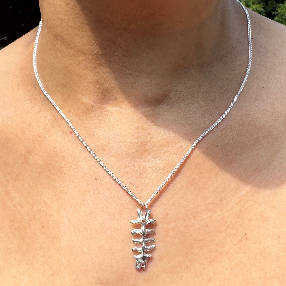 A person wears a Soufana Attia Baraka Pendant with a spine-shaped design, displayed on bare skin and paired with a magenta off-shoulder top, reflecting traditions of protection.