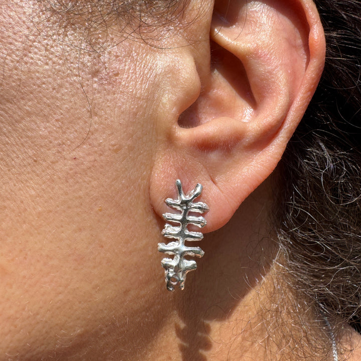 A close-up of a person's ear showcases the Baraka Earrings by Soufana Attia, silver fish skeleton earrings inspired by Arab symbols of protection. The earring's shadow falls on the skin, with the background softly blurred.
