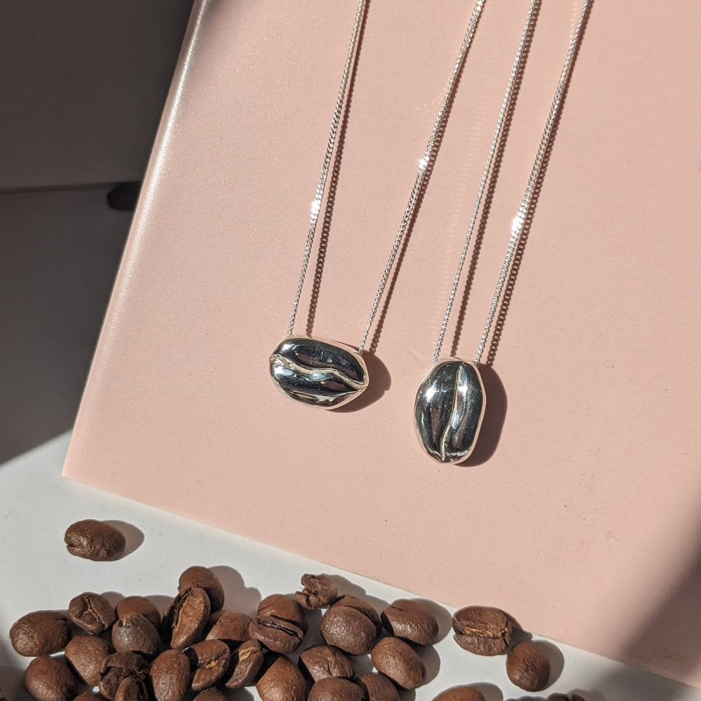 The Barista Bean Coffee Necklace Portrait by Becca Calmont features two recycled silver bean-shaped pendants on delicate chains, displayed on a pink surface with scattered coffee beans—perfect for coffee-loving jewellery enthusiasts.