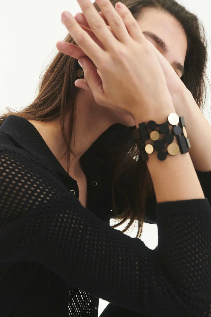 A woman with long brown hair covers part of her face with her hands. She is wearing a black netted top and the Bauhaus Bracelet by Iskin Sisters, featuring chunky black and gold circular pieces. The background is plain white.