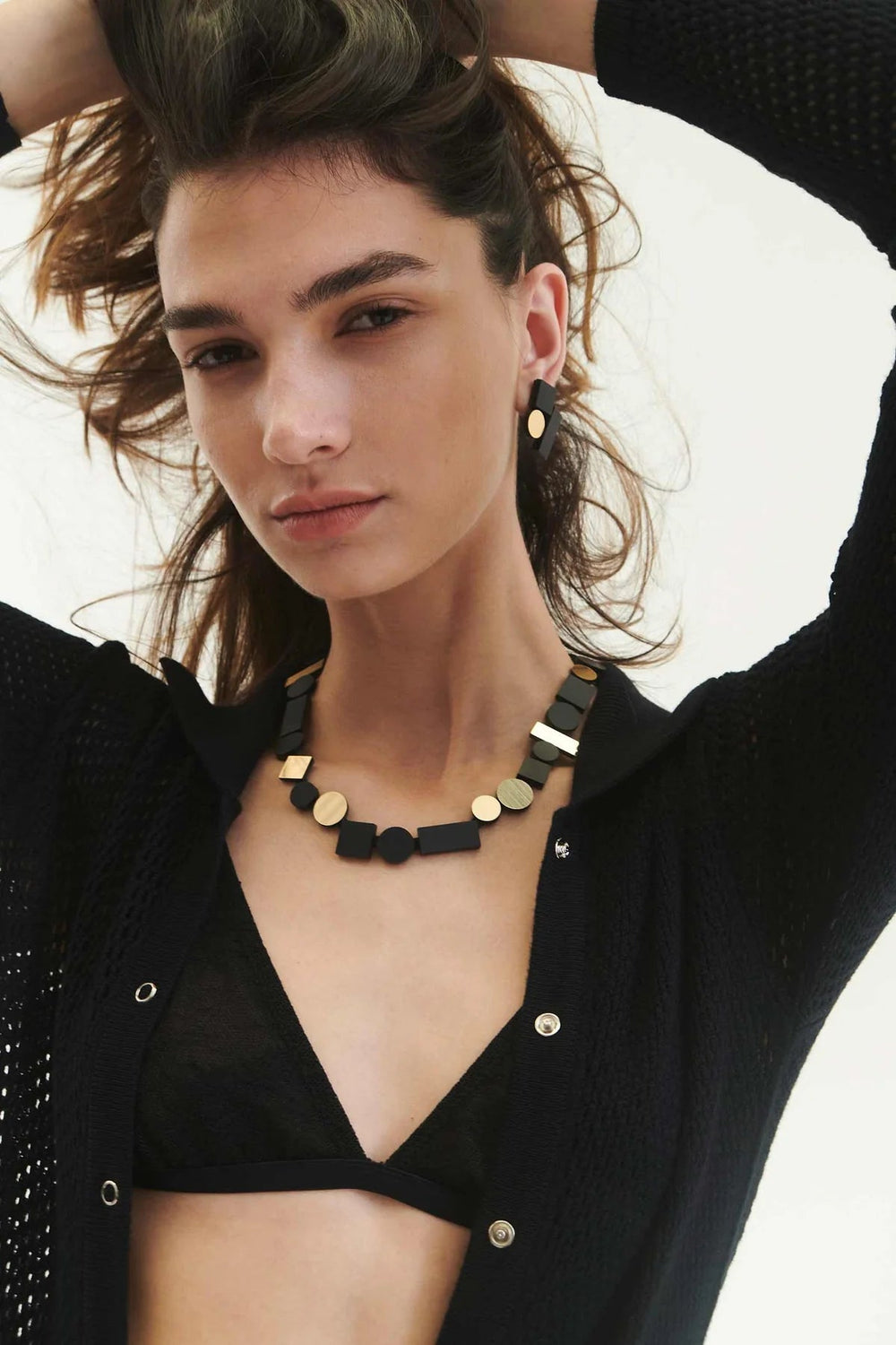 A woman with long brown hair is posing with her hands in her hair. She is wearing a black cardigan over a black bra top, complemented by the Bauhaus Black & Gold Necklace and matching earrings by Iskin Sisters. She has a neutral expression on her face and is looking at the camera.