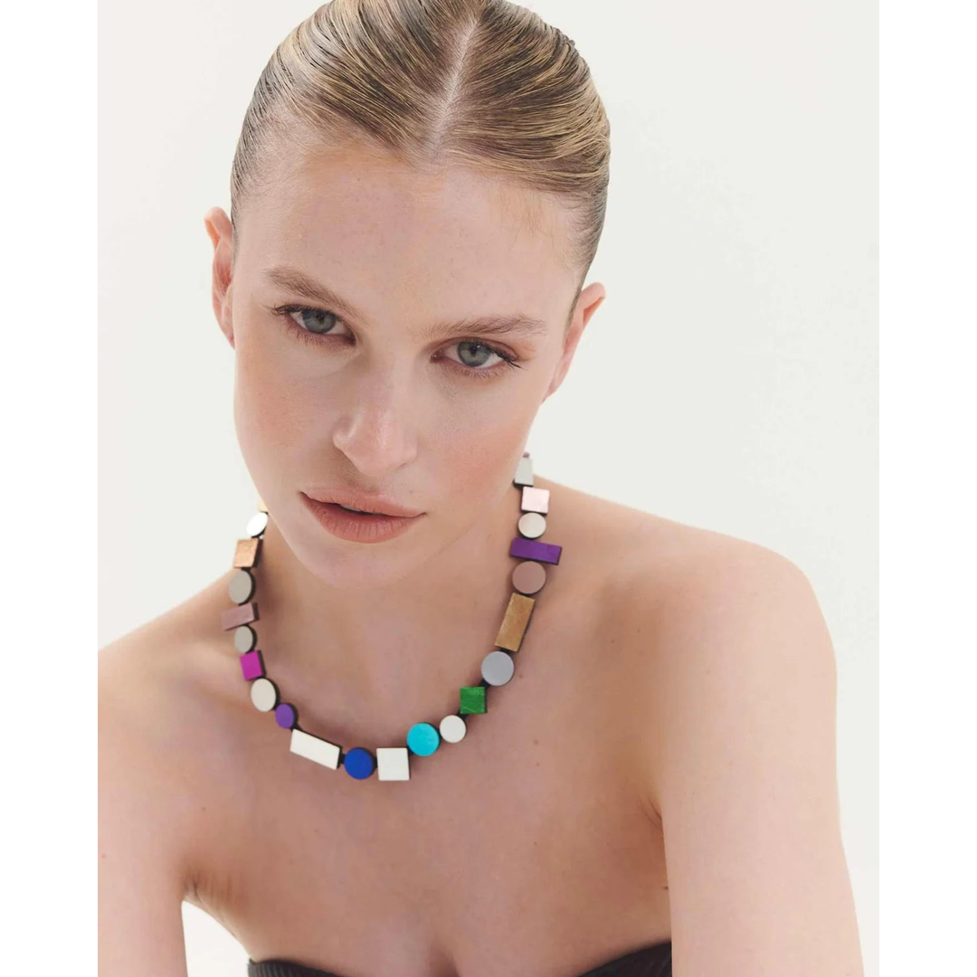 A woman with slicked-back hair wears a strapless dark top and the Iskin Sisters Bauhaus Rainbow Necklace, a modern statement piece featuring colorful geometric shapes in purple, blue, white, and gold against a light background.