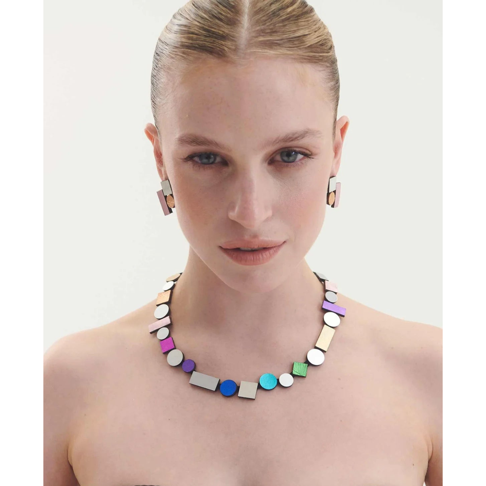 A woman with slicked-back hair wears geometric earrings and the Iskin Sisters Bauhaus Rainbow Necklace, whose colorful circles and rectangles make a bold modern statement against a plain light background.