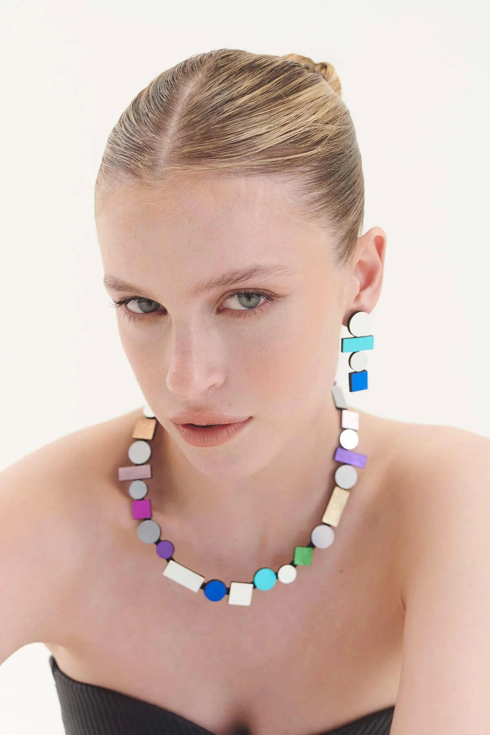 A woman with a slicked-back hairstyle is wearing large Bauhaus earrings in various colors from Iskin Sisters, featuring a combination of square and circular shapes in vibrant hues. She is adorned with a matching geometric necklace and clad in a black, strapless top, looking directly at the camera against a plain white background.