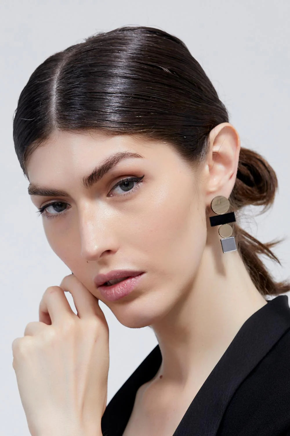 A person with dark, slicked-back hair and light skin is wearing a black top. They are adorned with the Bauhaus Earrings Large in various colours from Iskin Sisters, featuring a mix of black and metallic square shapes. The person's hand is gently resting near their chin as they look directly at the camera.