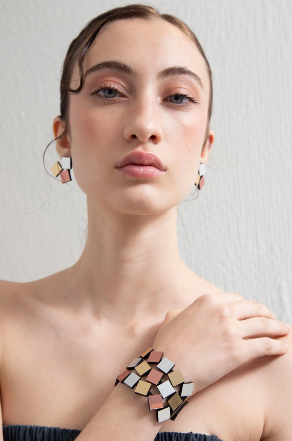 A close-up of a model wearing geometric mosaic earrings and a matching geometric bracelet. The jewelry features faceted tiles in silver, gold and copper colour, set against a neutral light grey background.