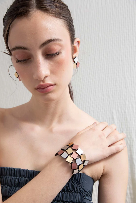 A close-up lifestyle shot of a model wearing a geometric mosaic cuff bracelet . The jewelry features a mix of gold, rose gold, silver squares, styled on a model wearing a dark strapless top against a light textured wall.