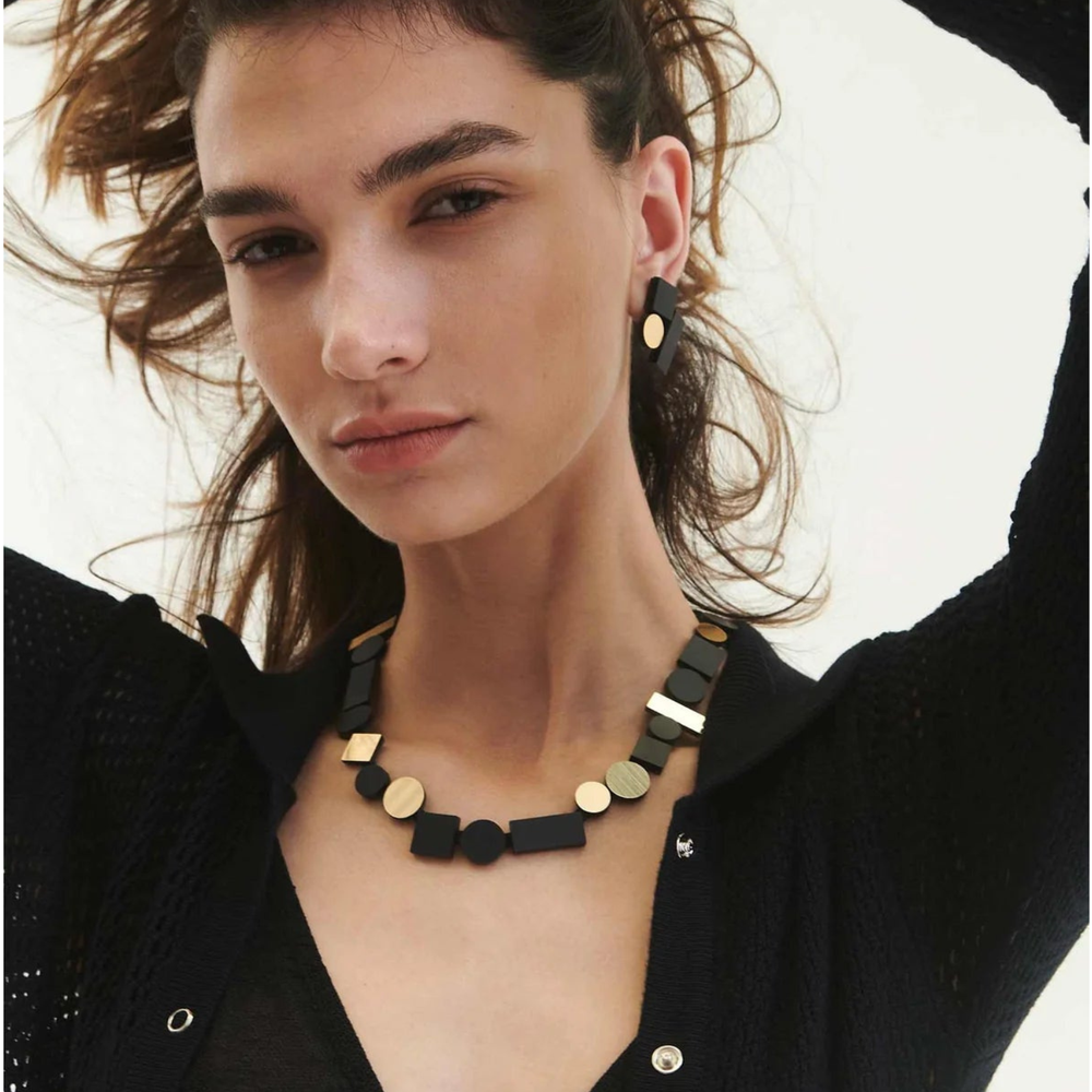 A woman with wavy brown hair wears the Iskin Sisters Bauhaus Black & Gold Necklace and matching earrings with a black textured cardigan, gazing confidently at the camera against a light background.
