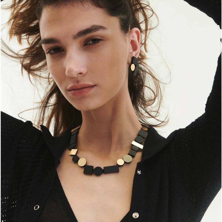 A woman with wavy brown hair wears the Iskin Sisters Bauhaus Black & Gold Necklace and matching earrings with a black textured cardigan, gazing confidently at the camera against a light background.