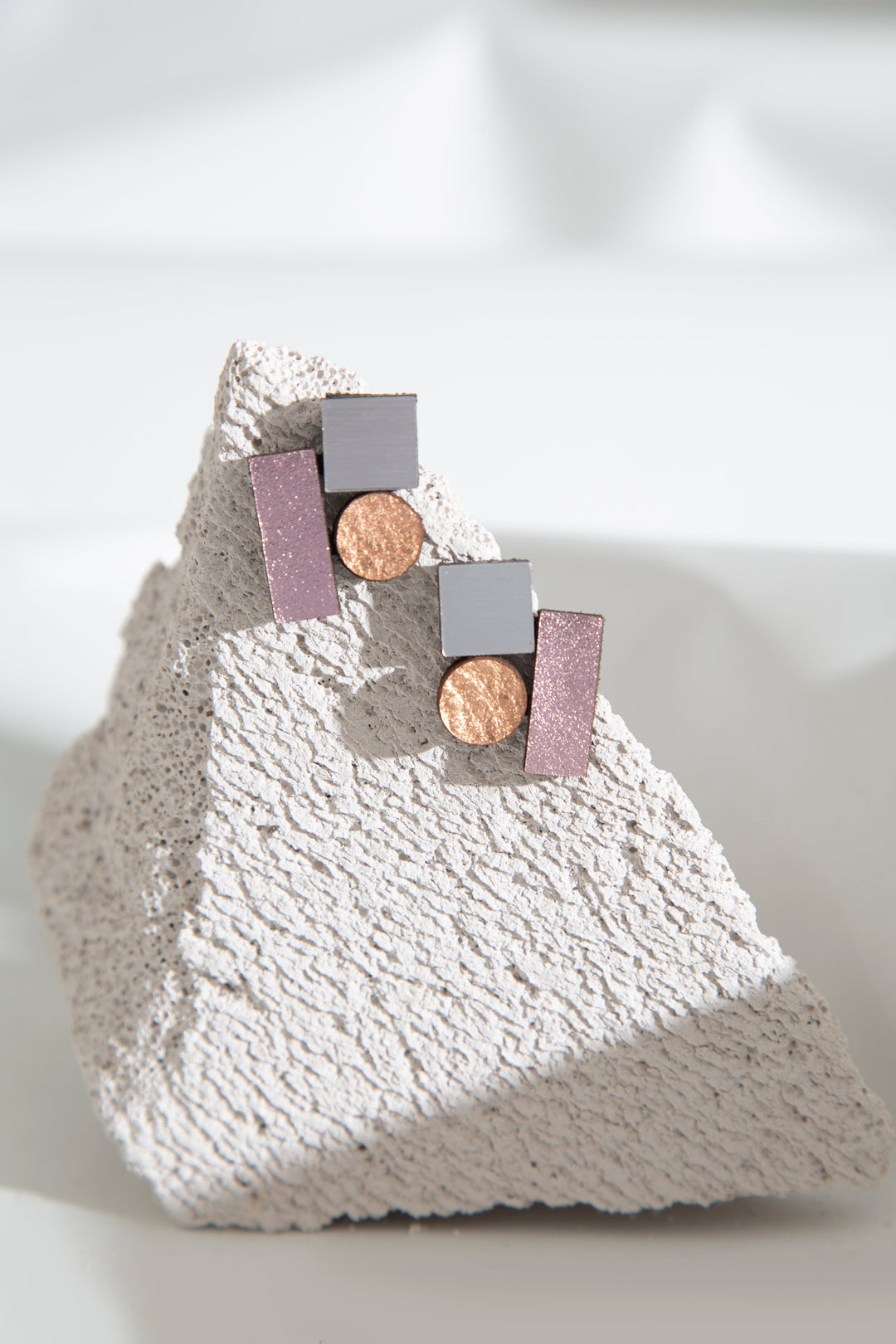 A pair of Bauhaus Earrings Small in various colours from Iskin Sisters is displayed on a textured stone. The earrings feature silver squares, pink rectangular bars, and copper circles arranged in a contemporary design. With exceptional materials and a featherlight structure, they are beautifully highlighted by the softly blurred background.