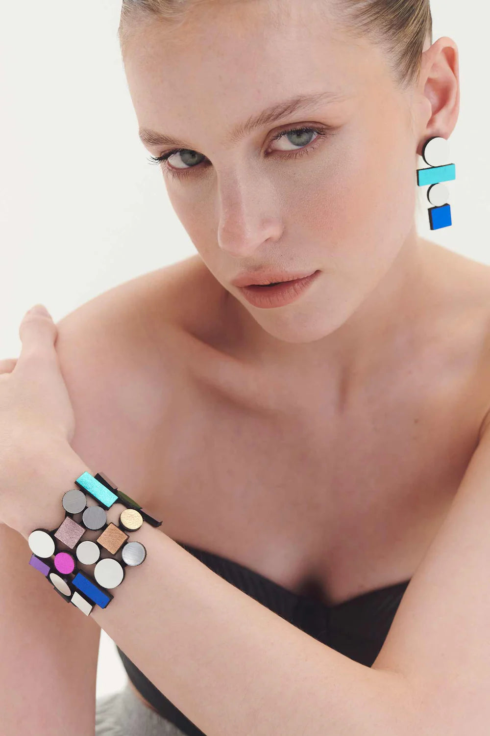 A person with a minimalist look wears geometrically shaped, colorful earrings and the Bauhaus Bracelet by Iskin Sisters. Dressed in a strapless black top, they look directly at the camera with a slight tilt of their head, showcasing the statement jewelry that exudes modernist craftsmanship.