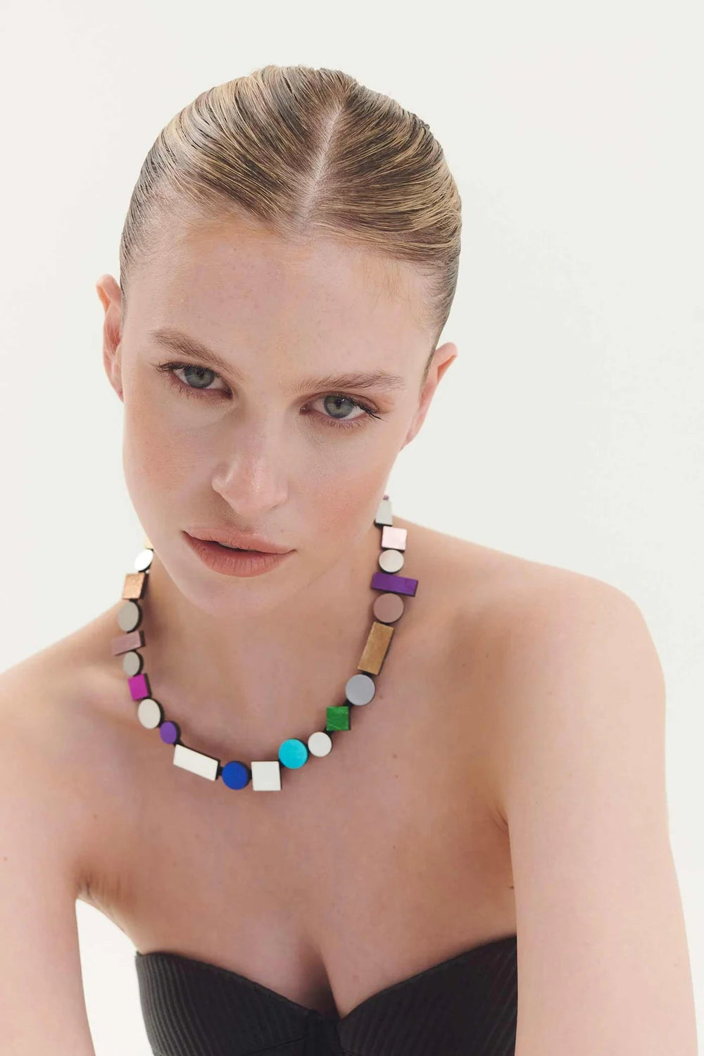 A woman with light skin and slicked-back blonde hair wears a strapless black top and the Bauhaus Rainbow Necklace from the Iskin Sisters. She looks directly at the camera with a neutral expression against a plain white background.