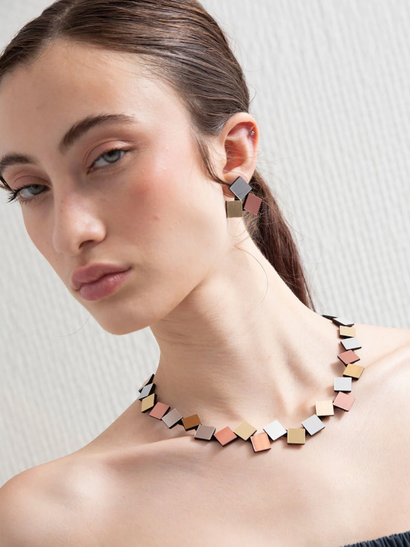 A model wears an Iskin Sisters Bauhaus Round Squares Necklace - Silver, Gold & Copper with matching earrings, featuring a series of small, square metal plates in silver, gold, and copper tones.