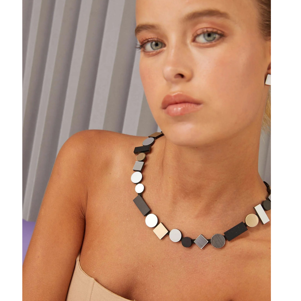 A woman with light eyes and nude makeup wears the Iskin Sisters Bauhaus Silver, Black & Gold Necklace and matching earrings. She pairs them with a strapless beige top against a neutral pleated background.