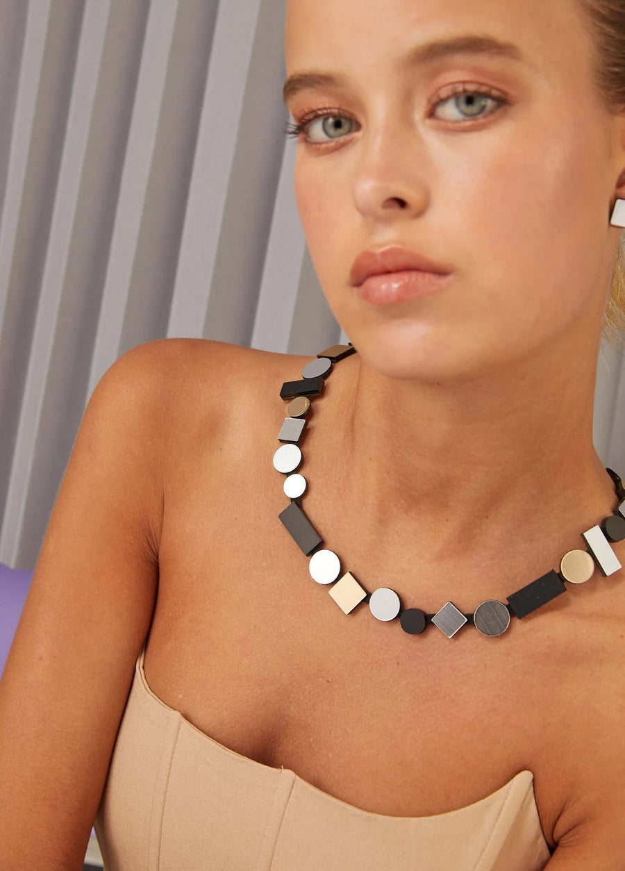 A woman with a light complexion and light hair is seen from her shoulders up, wearing a strapless beige top and a chunky statement necklace from the Iskin Sisters' collection. The necklace, called the Bauhaus Silver, Black & Gold Necklace, features black and metallic square and circular shapes. The background showcases vertical, light-colored paneling.