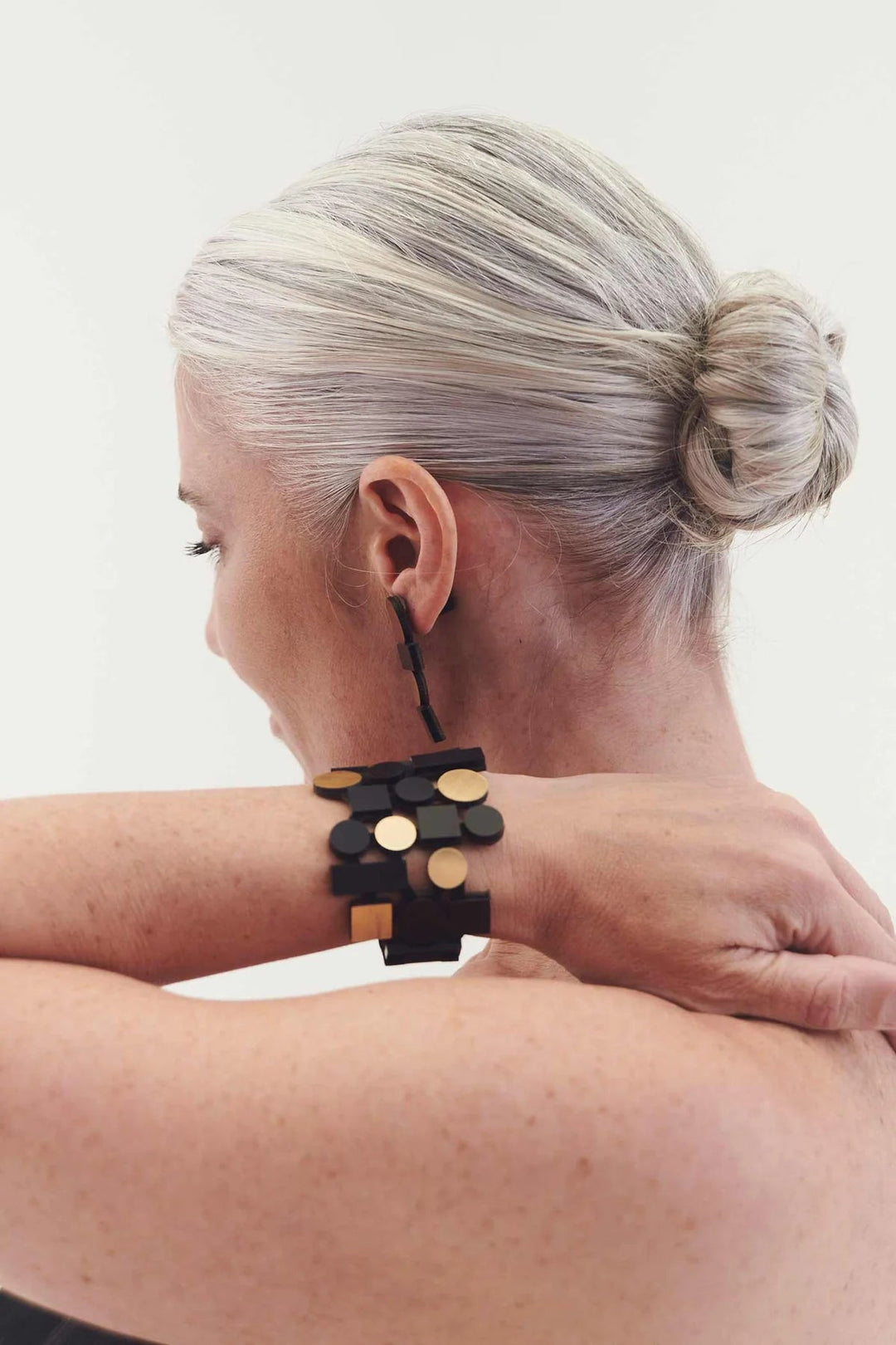 A person with gray hair styled in a low bun wears a black earring with geometric shapes and the Bauhaus Bracelet by Iskin Sisters, featuring black and gold elements. The person's back is turned, and their hand is placed on their shoulder, displaying the jewelry prominently.