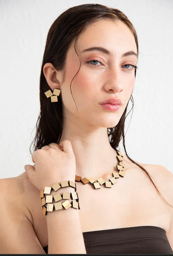 A woman with wet dark hair models a matching geometric gold-tone jewelry set, including a chunky necklace, matching earrings, and a wide cuff bracelet, against a plain light background.