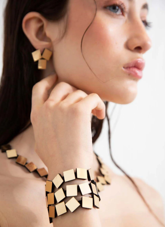 A close-up lifestyle shot of a model wearing a matching geometric jewelry set consisting of a gold-toned cube-link necklace, matching cube-link bracelet, and cube-link earring.