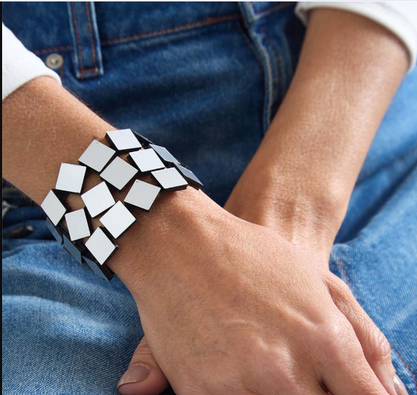 A close-up of a person's wrist wearing a geometric bracelet made of silver squares, with blue jeans and a white shirt visible in the background.