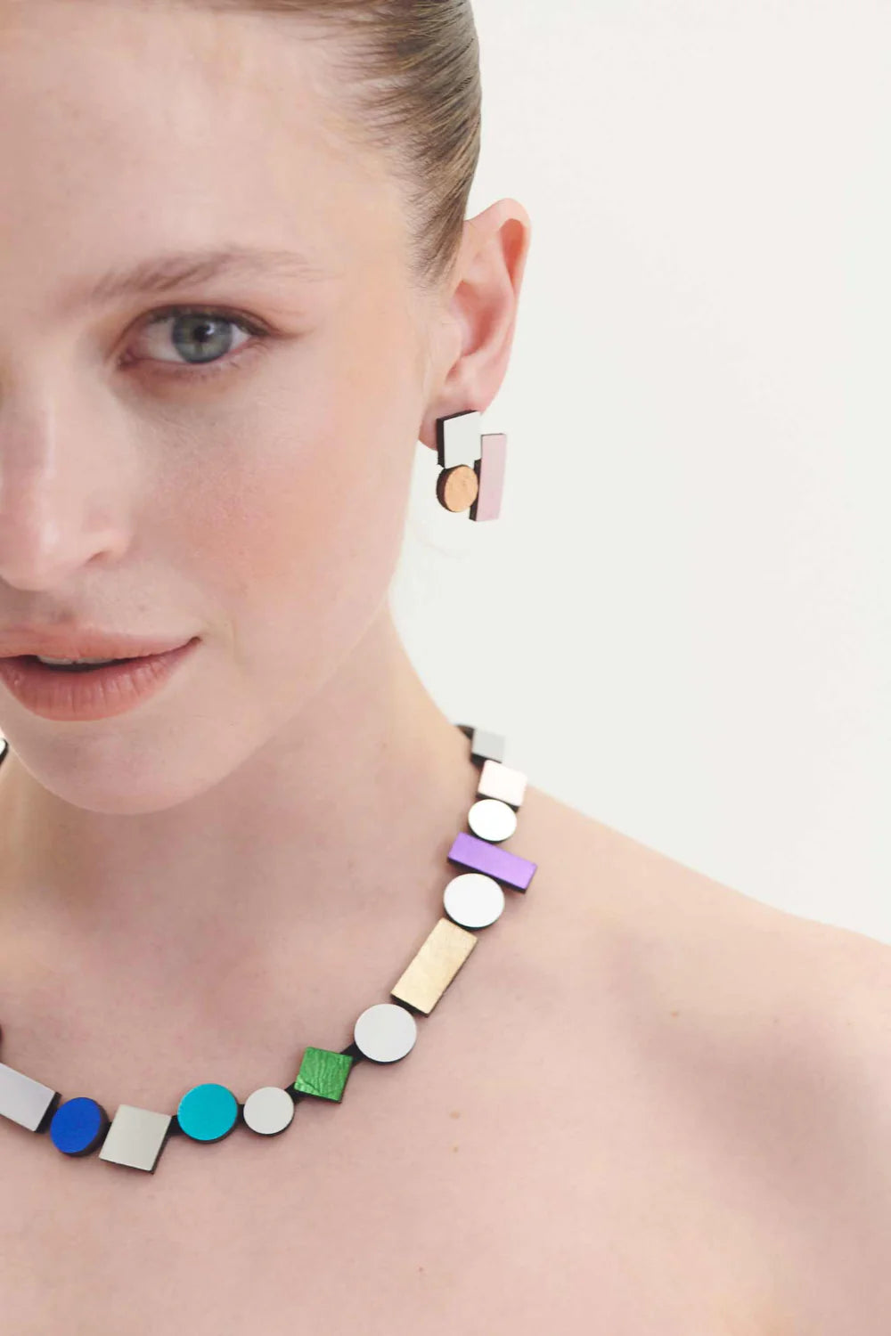 A woman with fair skin and light brown hair slicked back is wearing a modern, geometric necklace and matching Bauhaus Earrings Small in various colours from Iskin Sisters, featuring various colorful and metallic shapes. The background is plain white.