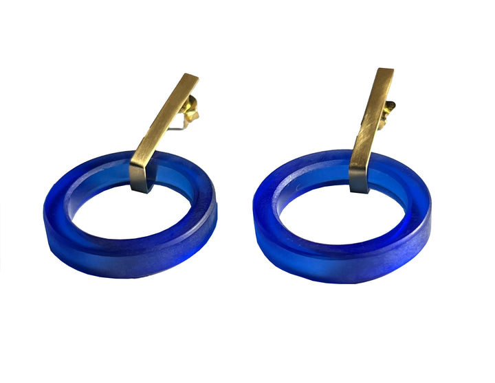 A pair of modern Soul Bottle Long Circle Earrings from Beira by Irene Reyes featuring blue, circular, translucent hoops made from recycled bottle glass with attached gold bars forming the earring hooks. The gold bars provide a sleek contrast to the vibrant blue hoops, creating a stylish and contemporary design.