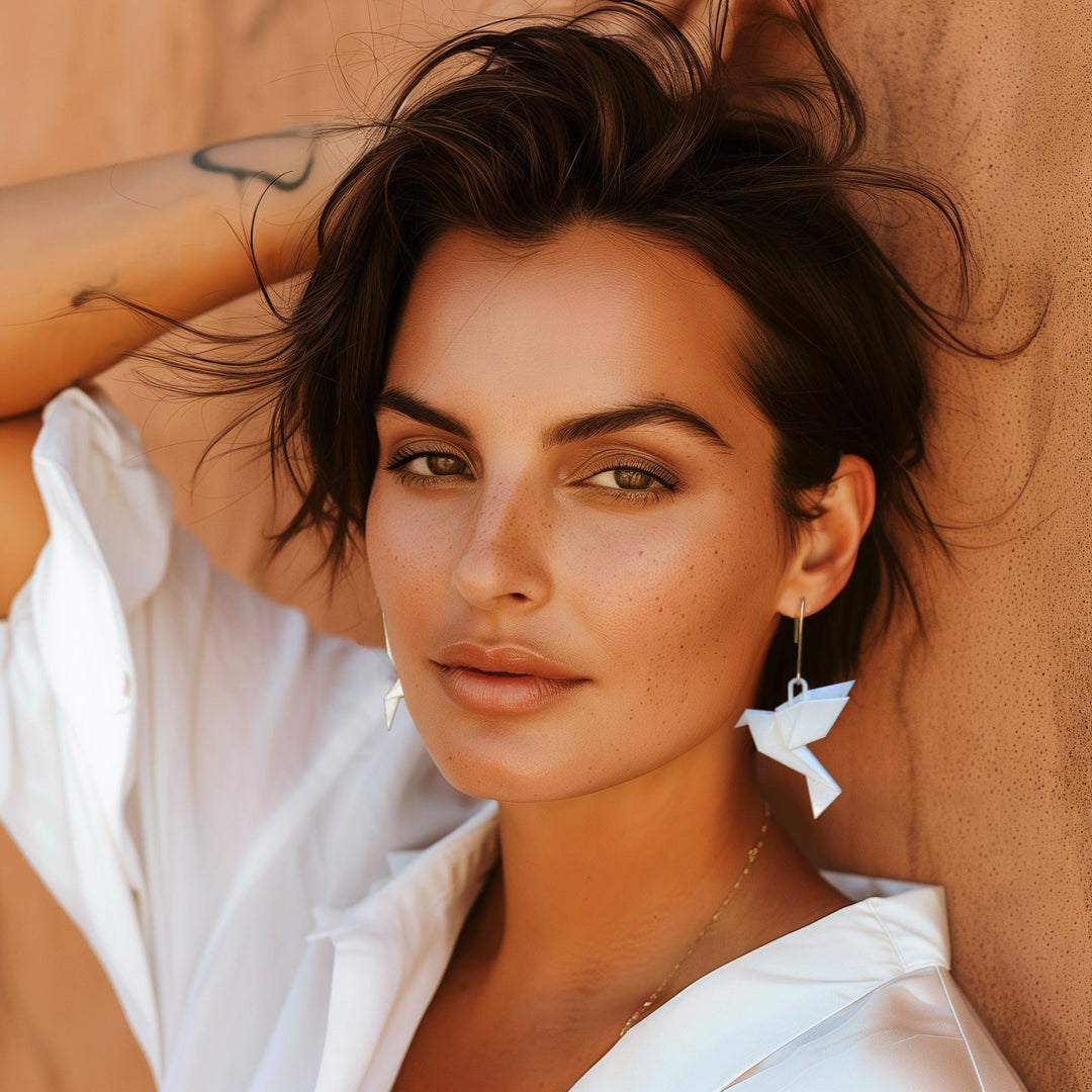 A woman with short dark hair and light makeup poses against a tan wall, wearing a white shirt and Tochu-De Origami Bird Aubergine Earrings. With one arm raised, she displays a relaxed expression and a forearm tattoo.