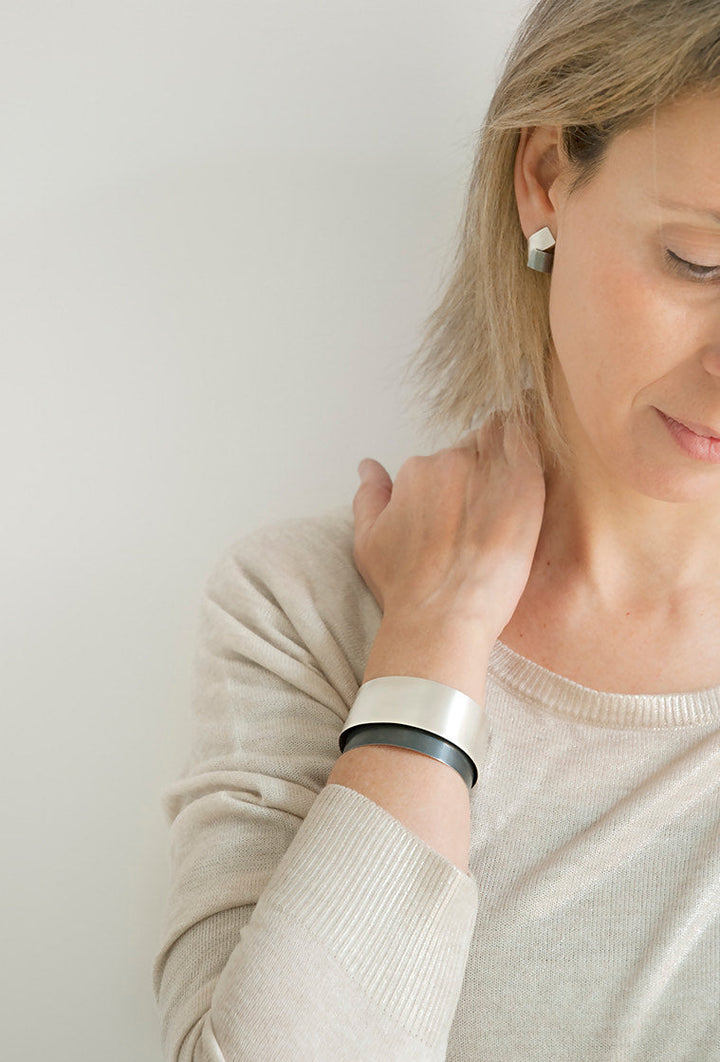 A person is wearing a beige sweater, silver geometric earrings, and a Bilbao Bracelet from MarCucurella featuring oxidized silver elements. The individual has light-colored hair and is looking downward with one hand resting on their neck. The background is white and minimalistic.