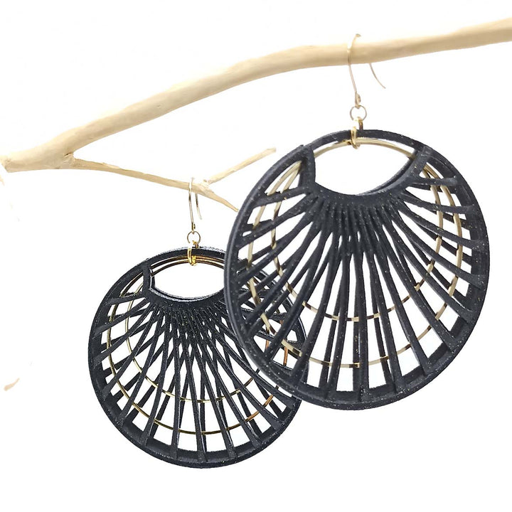 Two black circular earrings, made from powder-coated 3D printed nylon and featuring a geometric, fan-like cutout pattern, hang from a light-colored branch. Golden hooks attach them to the branch. The white background accentuates the dark earrings and light branch. Remember jewellery care for lasting beauty with your **Black Waterlily Earrings by Tochu-De**.