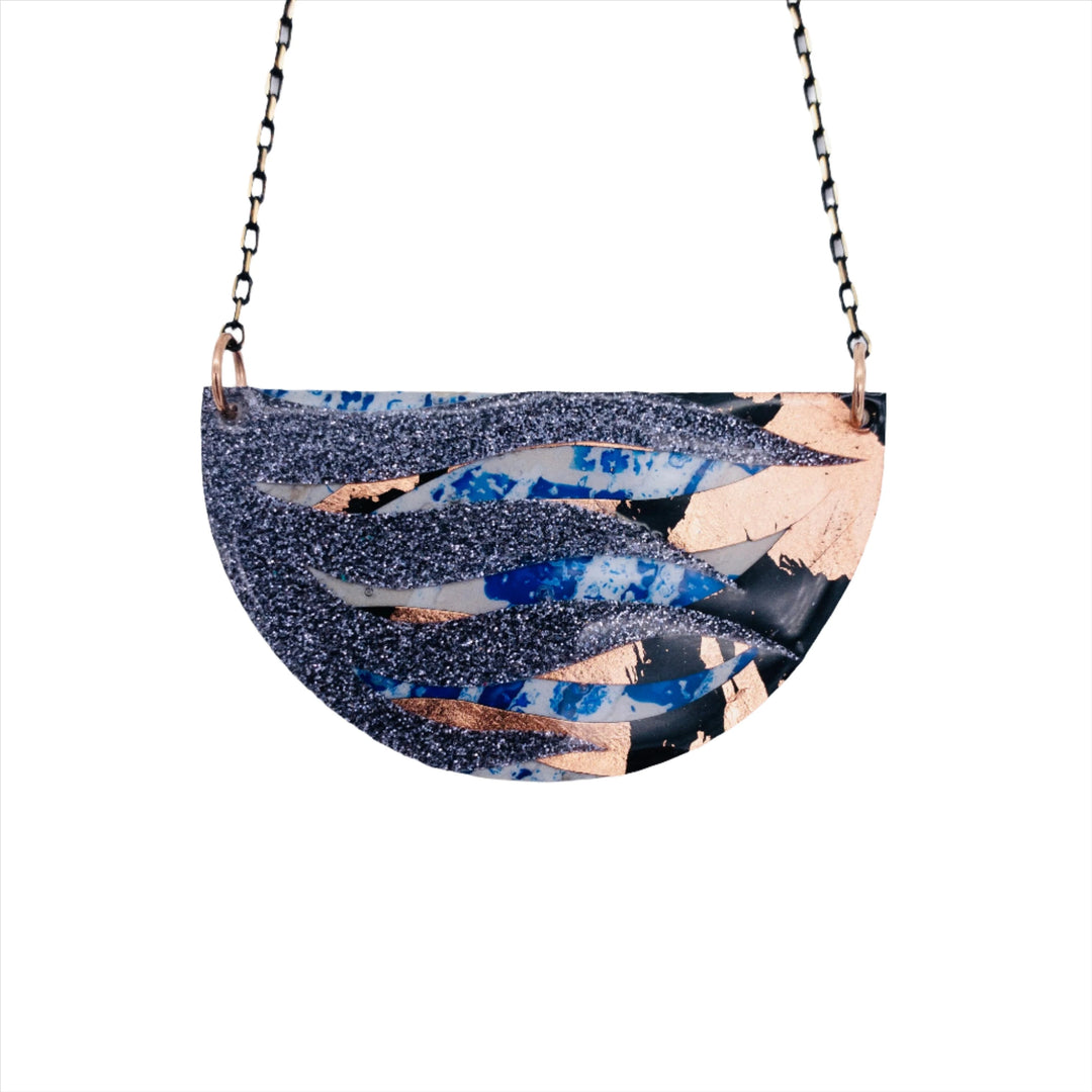 Introducing the Bláth Batik Textile Necklace by Rothlú, featuring a semi-circular pendant with an abstract design. This unique piece showcases a stunning mix of glittery dark blue, metallic rose gold, and blue and black splashes that create a flowing wave-like pattern. The high gloss eco resin finish enhances its elegance as the pendant is suspended from two rings at the top corners.