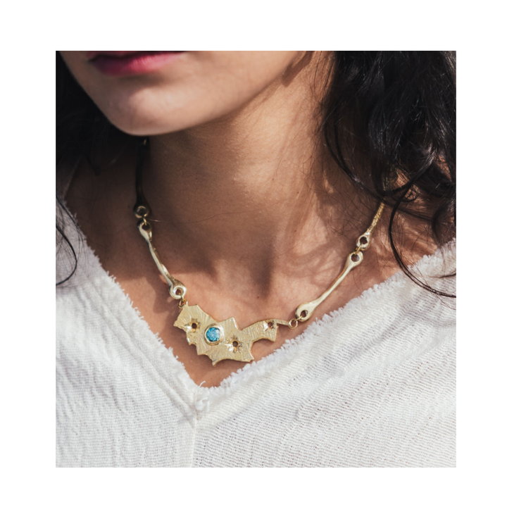 A woman wears a white textured top and the Madeleine Blaine Blaze Necklace, a unique gold piece with an abstract, jagged design featuring a blue gemstone, paired with sculptural earrings for an effortlessly artistic statement.