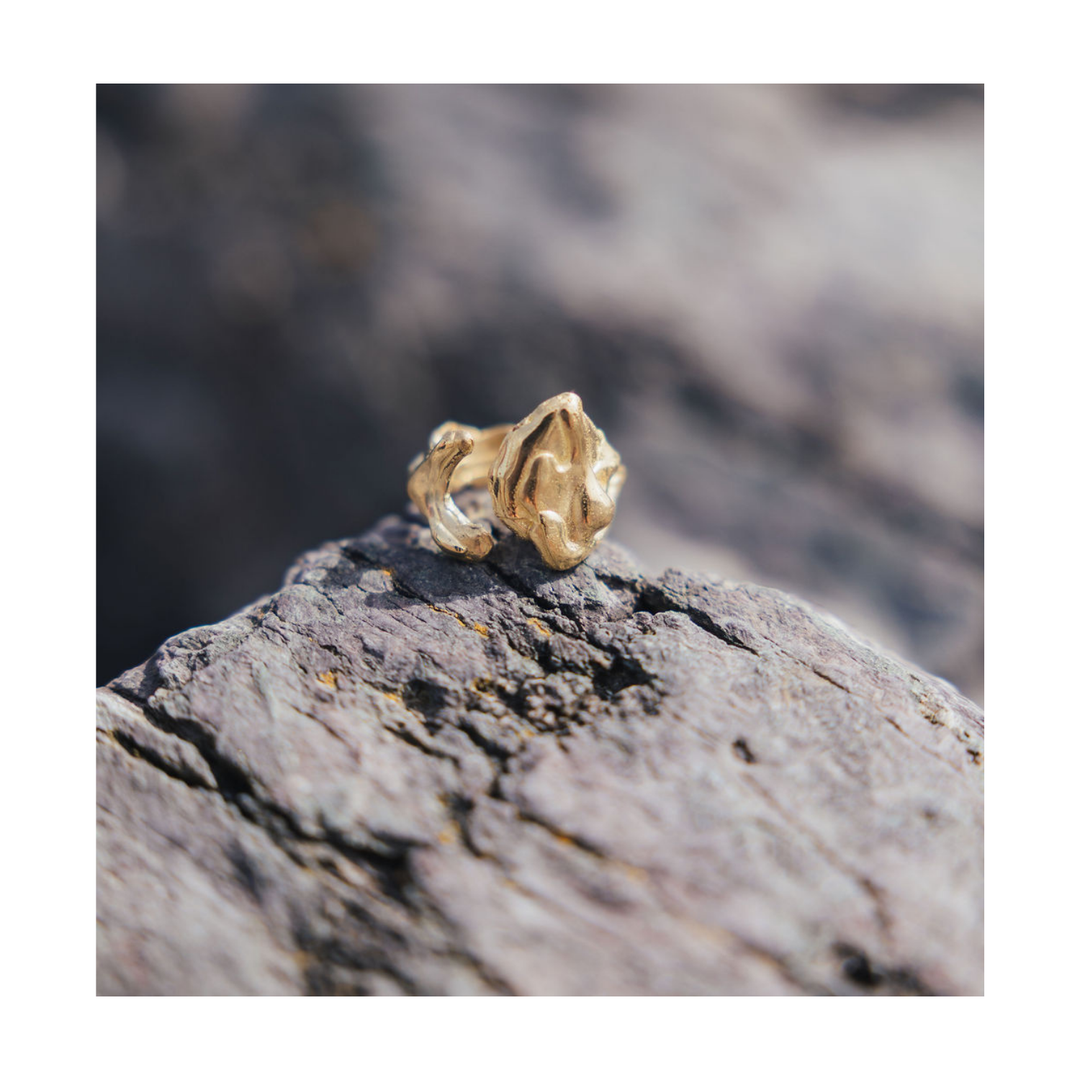 A textured gold ring with a sculptural, organic shape resting on a rough gray rock outdoors.