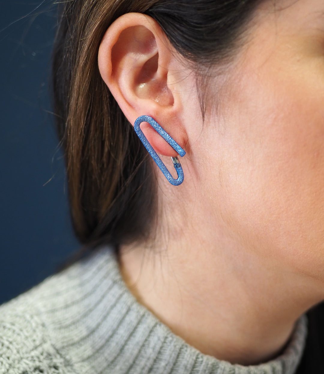 A person with long brown hair is shown from the side, focusing on their ear, which is adorned with a handcrafted earring shaped like O Silver Earrings with Blue Ceramic Coating by Theodora D. They are wearing a gray knit sweater, and the background is a solid dark blue.
