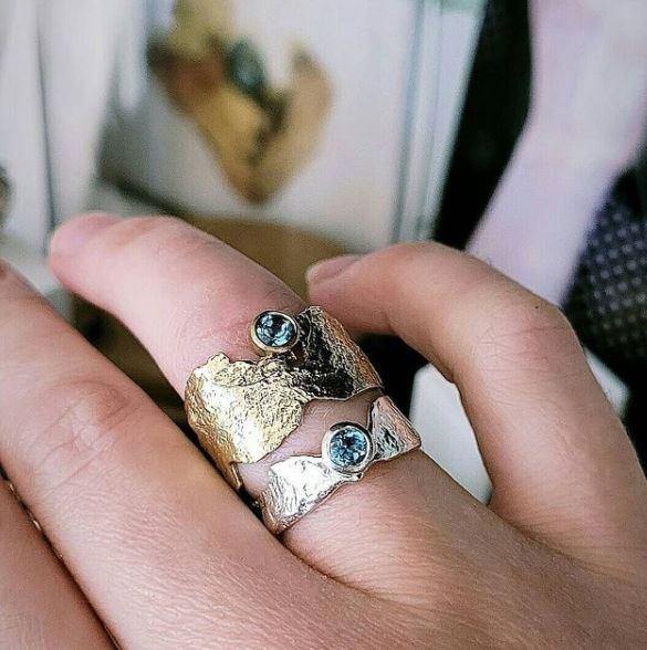 Close-up of a hand wearing two intricately designed silver and gold Faerie Tale Ring In Blue Topaz by Caroline Stokesberry-lee, each featuring a Blue Topaz gemstone set in the center using reticulation technique. The background is blurred, highlighting the ethereal Faerie Tale Ring In Blue Topaz by Caroline Stokesberry-lee on the hand.