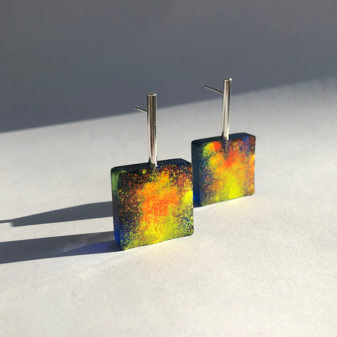 A pair of square-shaped, handmade earrings with a silver post and vibrant, abstract design. These Graffiti Block Earrings by Angela O'Keefe feature a blend of bright yellow, orange, and blue colors resembling a cosmic or nebula pattern. They stand upright on a light-colored surface, exuding artistic flair.