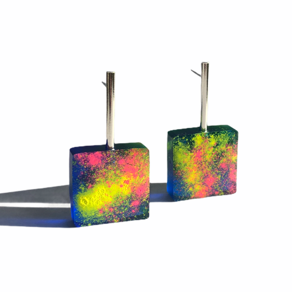 A pair of square, handmade resin earrings with vibrant neon splashes of yellow, pink, and green on a dark background. Dubbed the Graffiti Block Earrings by Angela O'Keefe, they have silver hooks for wearing and are positioned upright on a white surface with a slight shadow cast to the left.