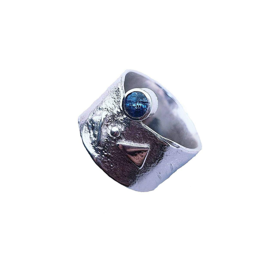 The Blue Topaz Guardian Ring by Caroline Stokesberry-lee features a textured Sterling Silver band with an abstract face design, Blue Topaz gemstone eye, silver accent, and raised triangle—a bold statement piece.