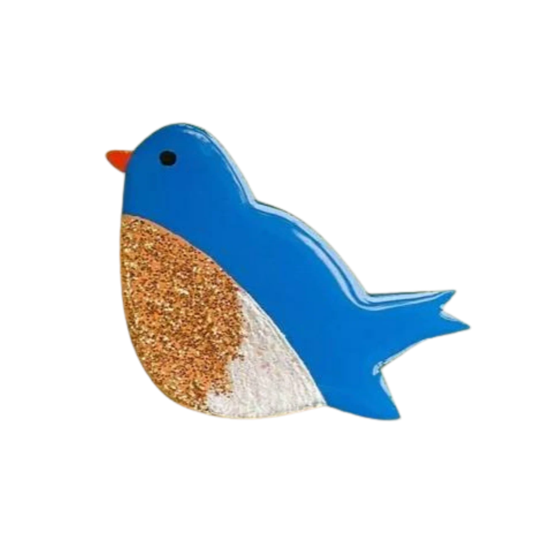 A colorful art depiction of a handpainted wooden Bluebird Pin Brooch by Naoi. The bird's head and tail are painted bright blue, its beak is orange, and it features a sand-textured body section with a speckled and lightly glittery appearance. The bird has one small black eye.