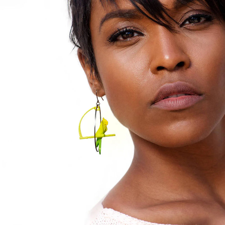 A person with short dark hair and smooth skin looks at the camera, wearing bold Tochu-De Citrus Origami Parrot Earrings featuring gold accents and geometric yellow-green patterns. The background is plain white.