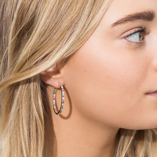 A close-up shows a woman with light skin and blonde hair tucked behind her ear, wearing Sif Jacobs Bovalino Hoop Earrings—large gold plated hoops adorned with small multicolored cubic zirconia gemstones.