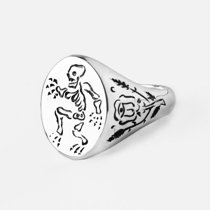 A Cosmic Boulevard Sterling Silver Memento Mori Signet Ring with an engraved skeleton on the circular face and a detailed rose design etched on the side. The white background highlights the intricate engravings, making this hand engraved piece a striking memento mori signet ring.